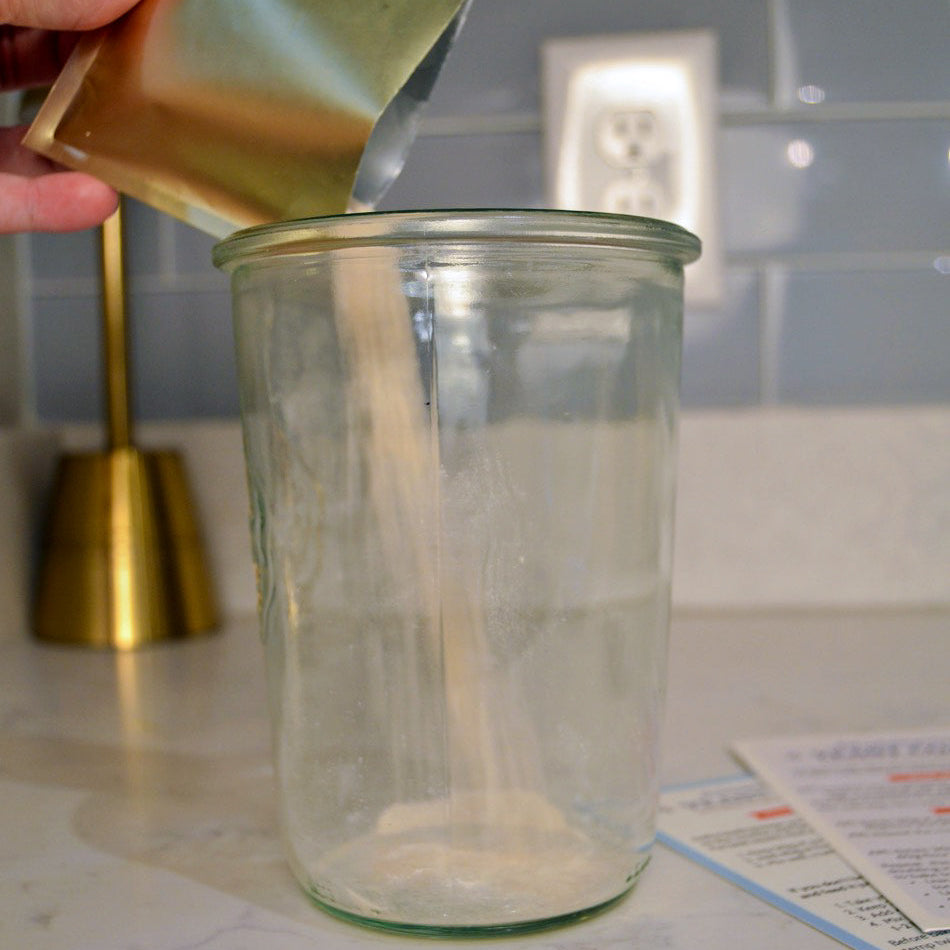 Hand pouring a green powder from a packet into a clear glass jar on a kitchen counter.
