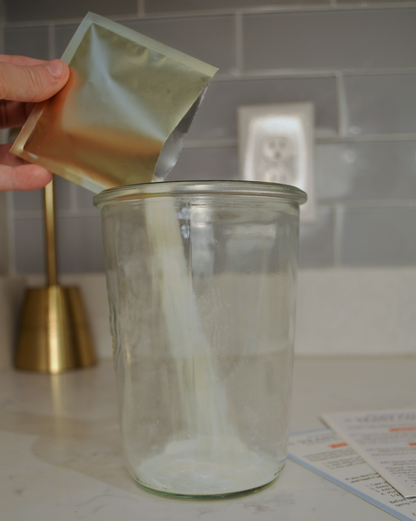 Hand pouring a white powder from a small container into a glass jar on a kitchen counter.